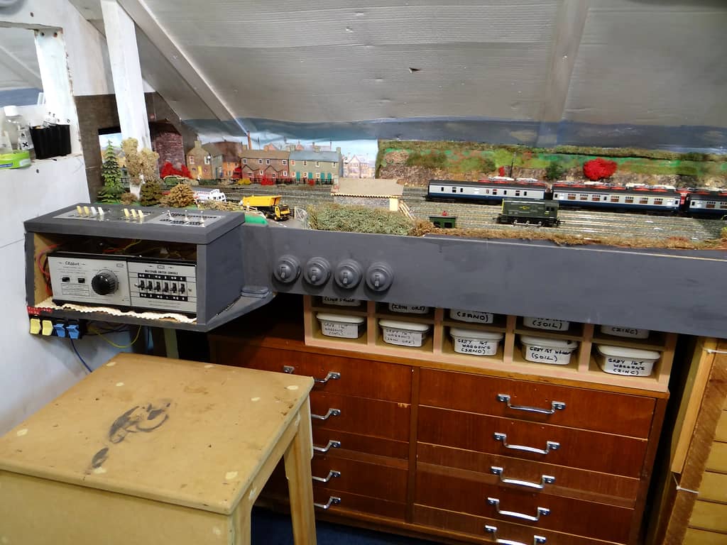 A grey control box with a power unit and a switch console hangs at the end of a baseboard, above tubs in pigeonholes and a dark wooden chest of drawers.