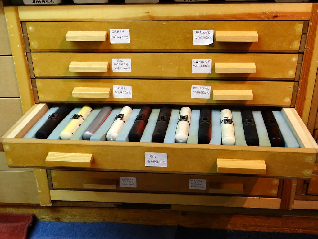 A wooden chest of drawers with handwritten labels, one drawer pulled out to show eleven tank wagons lying in blue foam slots.