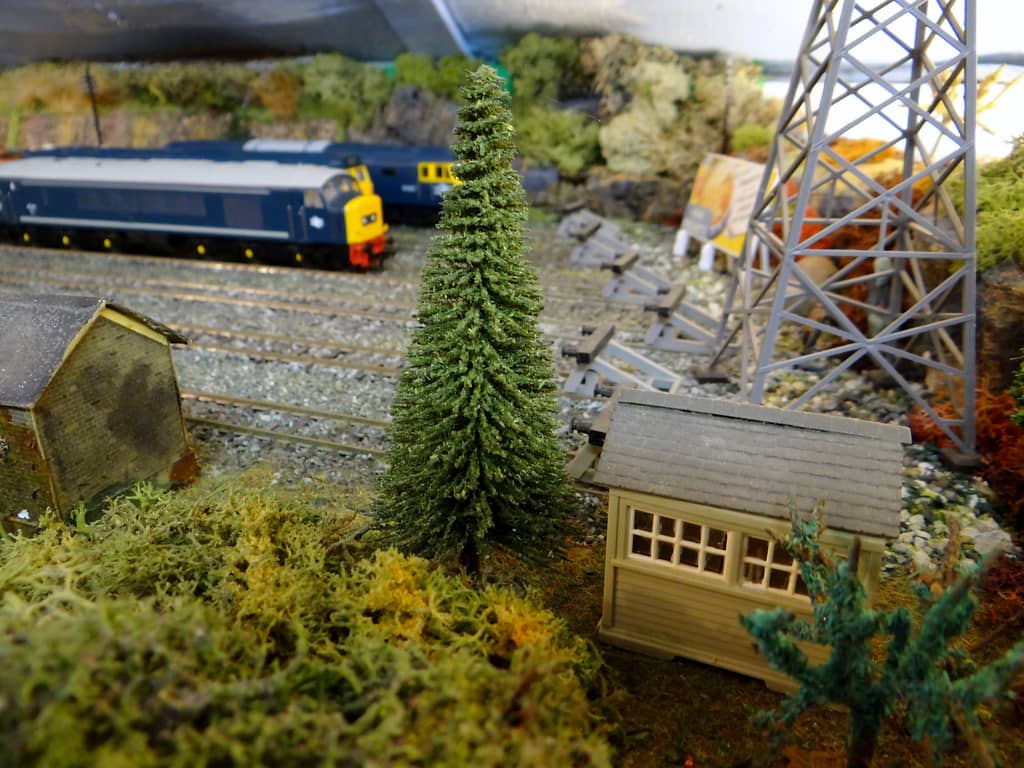 Low view of a cream wooden cabin with small-paned windows and a tall model fir tree, with grey buffer stops, a lattice pylon and blue diesels beyond.