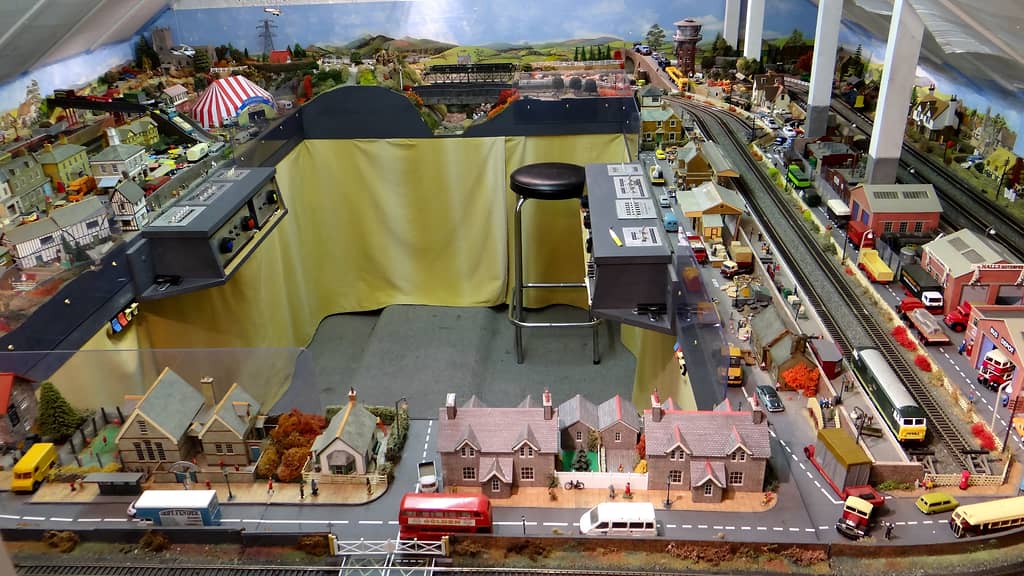 A model railway layout seen from above, wrapped round a well lined with yellow curtains, with a black stool, two control panels and a painted sky behind.