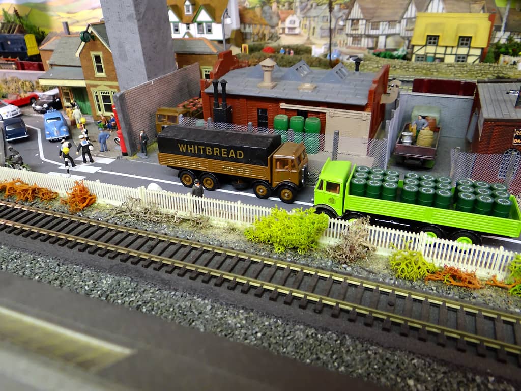 A brown lorry with a black “WHITBREAD” sheet and a lime-green lorry loaded with green barrels, parked by a picket fence beside a railway line, a brewery behind.