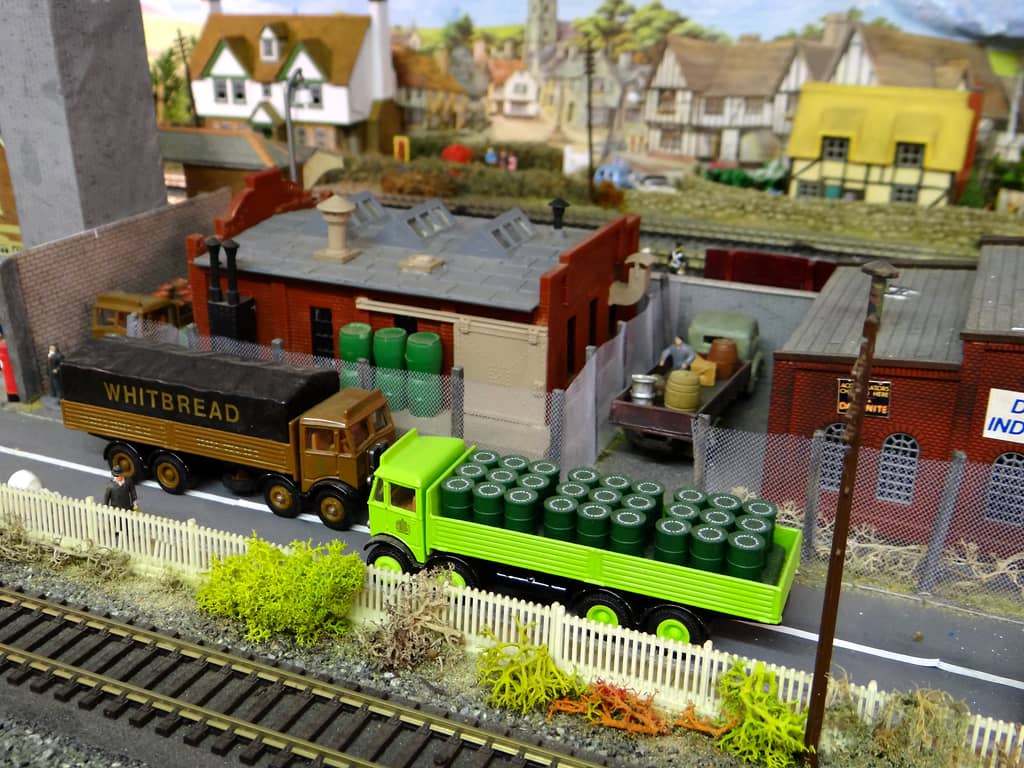 Two lorries parked beside a railway line, a brown Whitbread one with a black sheet and a lime-green one piled with green barrels, in front of a red-brick brewery.