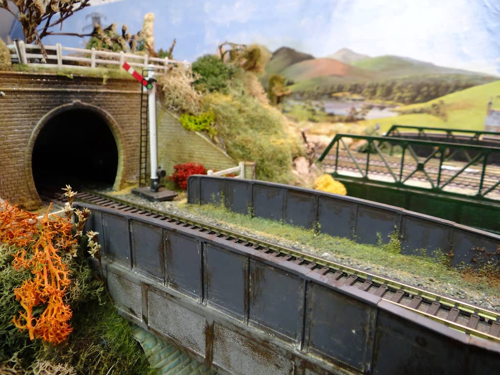 A brick railway tunnel mouth with a red and white semaphore signal beside it, a track crossing a grey girder bridge, and a green lattice bridge and painted hills beyond.