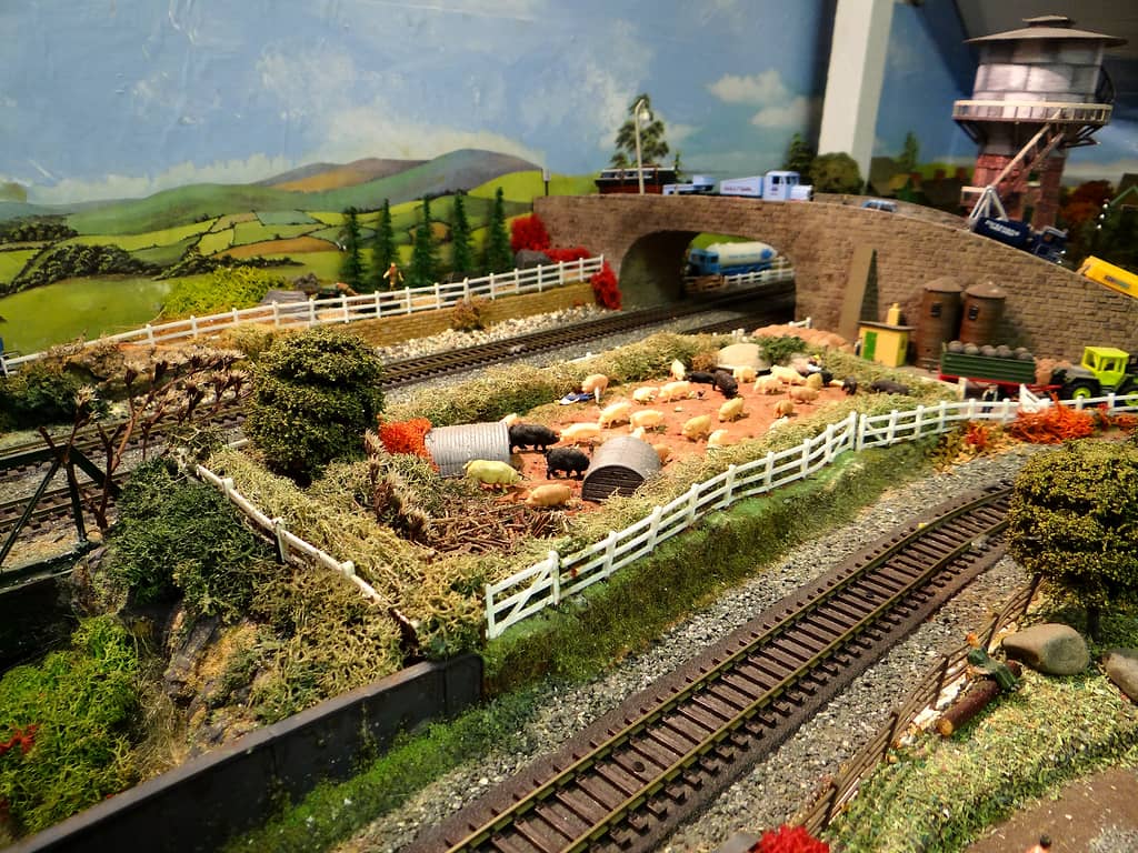 A fenced field of pink and black model pigs with grey corrugated shelters beside railway tracks, with a stone road bridge carrying lorries and a water tower behind.