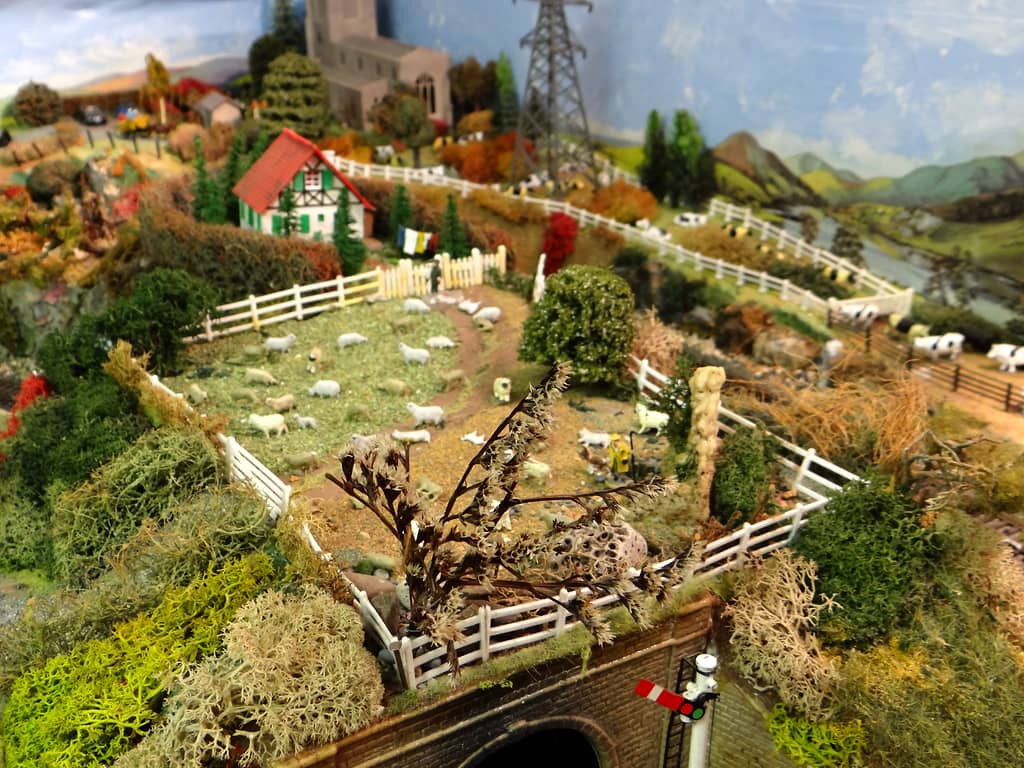 A sloping model field of white sheep inside white fences above a brick tunnel with a signal, with a red-roofed chalet, a church, an electricity pylon and cattle beyond.