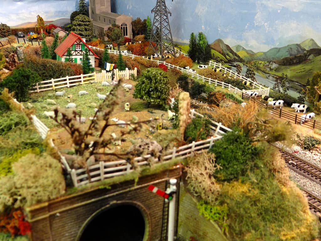 A model hillside with a stone church, an electricity pylon, a red-roofed chalet, sheep in fenced fields, cows by a gate and a brick tunnel with a signal at the bottom.