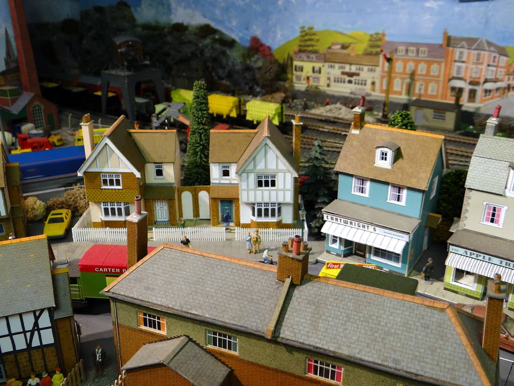 Model street of two detached houses behind white picket fences, a blue newsagents beside them, and a railway with yellow wagons and a crane beyond.
