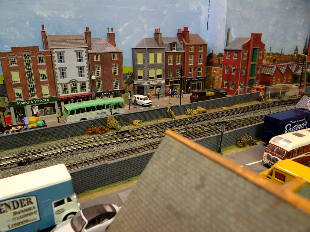 Model high street of brick and stone shops beyond a double railway line, with a pale green coach, a white van and a steam lorry in the road.