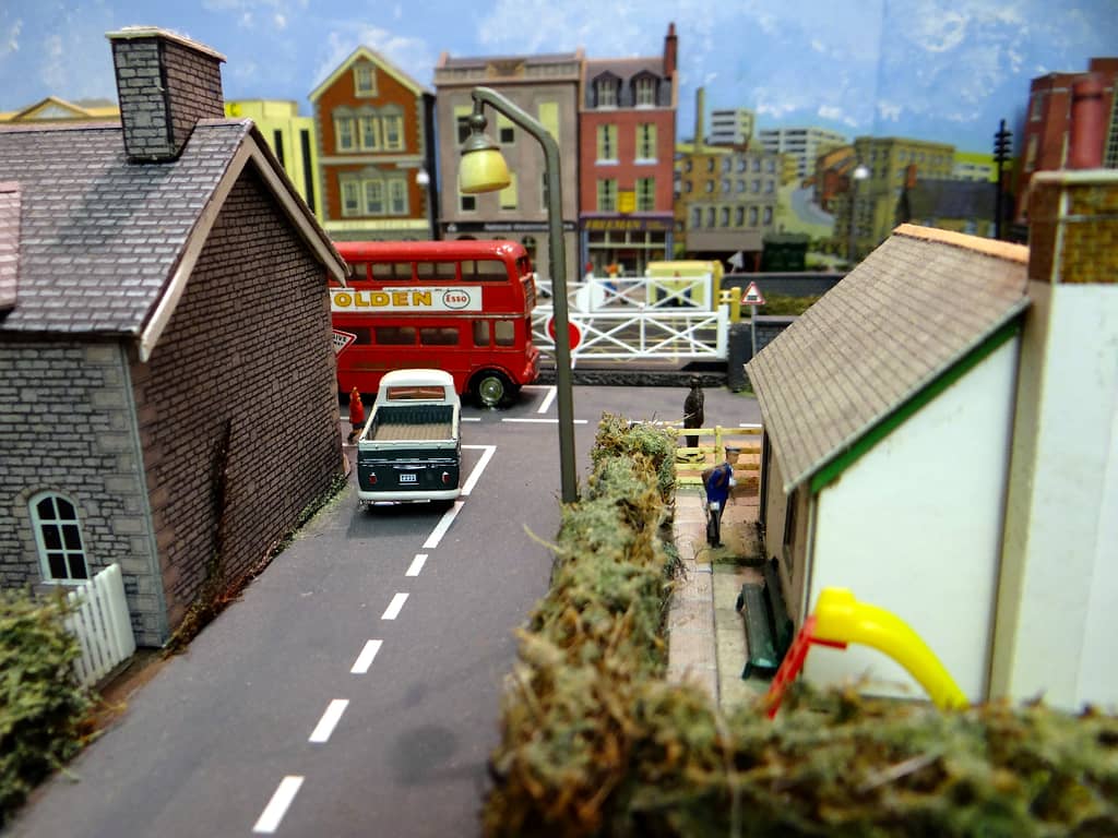 Model lane between two cottages leading to a road where a red double-decker bus passes white level crossing gates, with a green pickup truck waiting.