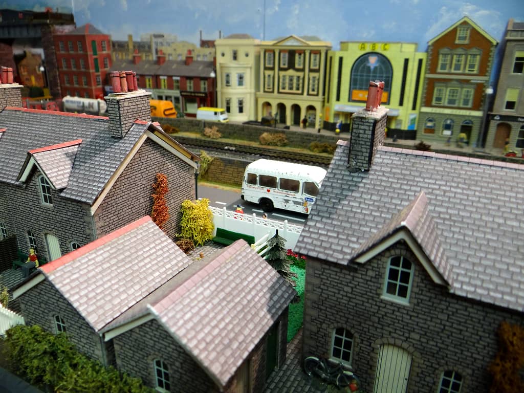 Grey stone cottage roofs in the foreground, a white minibus on the road beyond, and a railway with a yellow cinema and tall town buildings behind.