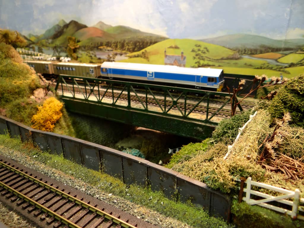 A silver-and-blue diesel pulls beige stone wagons across a green lattice girder bridge, with a grey plate girder bridge, track and a white farm gate in front and painted hills behind.