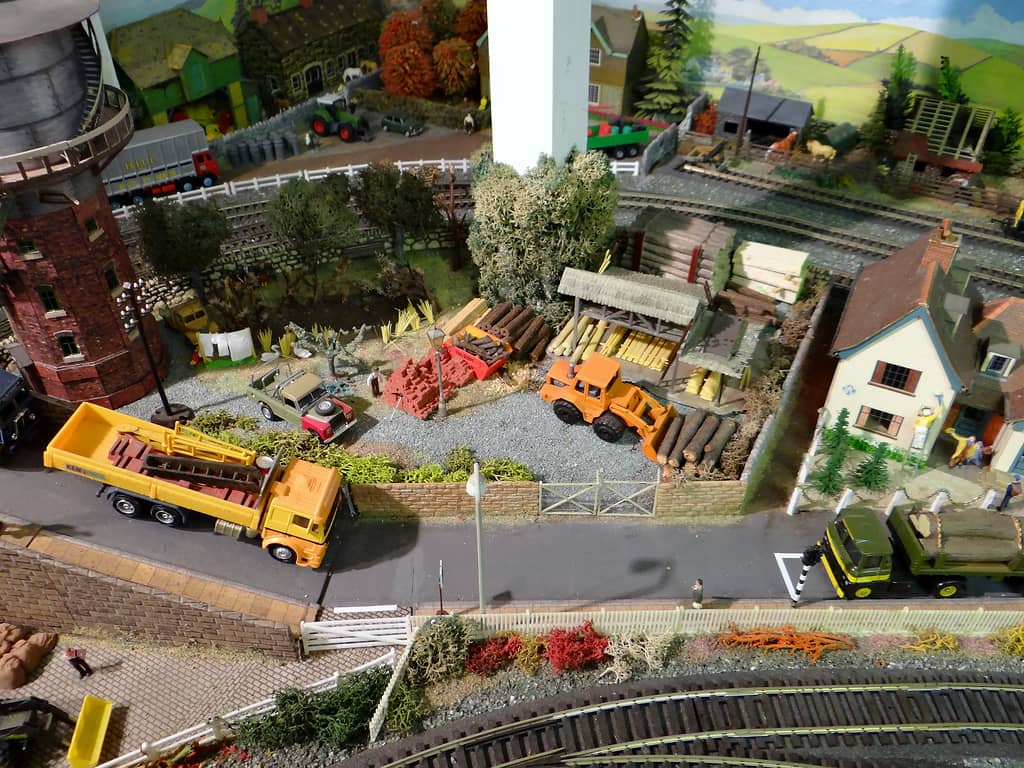 A model timber yard with stacked logs, a timber shed and an orange loading shovel, a brick water tower with a spiral stair at the left, a yellow lorry on the road and a cottage at the right.
