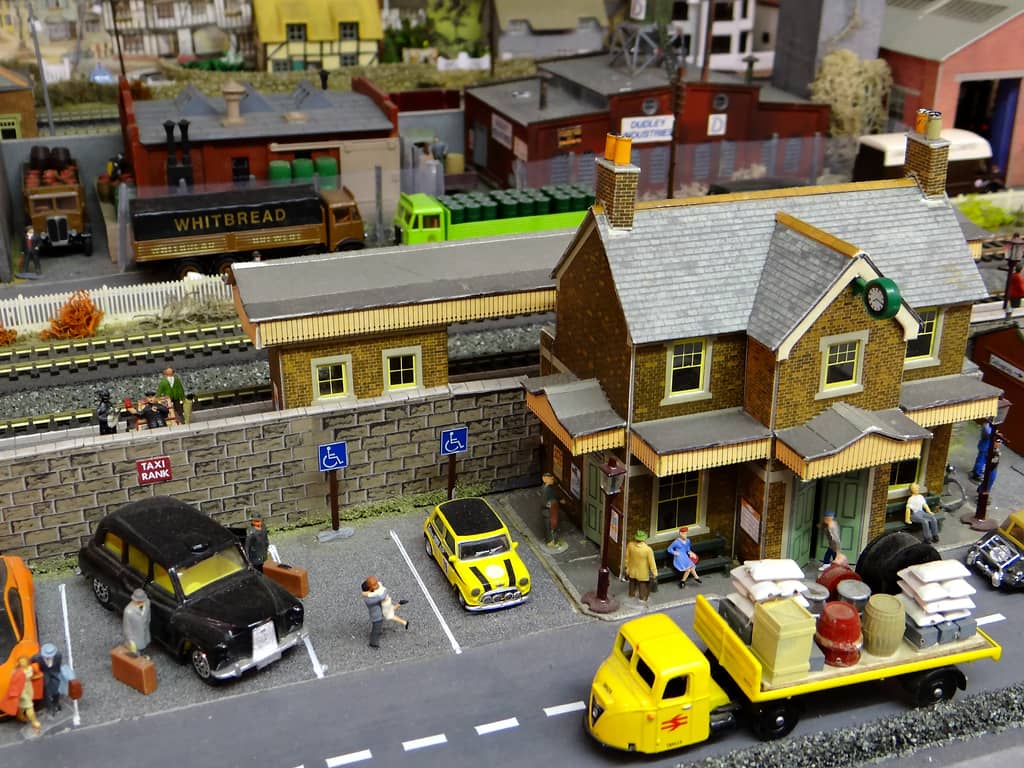 A yellow-brick model station with a slate roof and a clock on its gable, with a black taxi, a yellow Mini and figures in the car park, and a yellow three-wheeled lorry loaded with sacks and barrels in front.