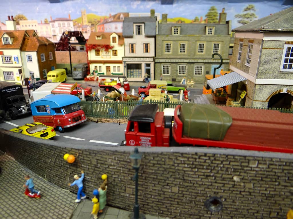 Model children playing ball against a stone wall, with a red London Brick lorry, a blue and red circus van and a row of shops round a market square behind.