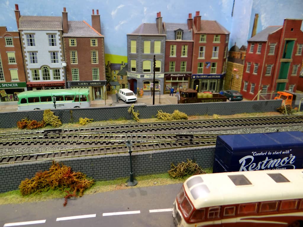 Model double railway line with a dark blue van and a cream and maroon coach in the foreground, and a row of shops, a pub and a red-brick warehouse beyond.