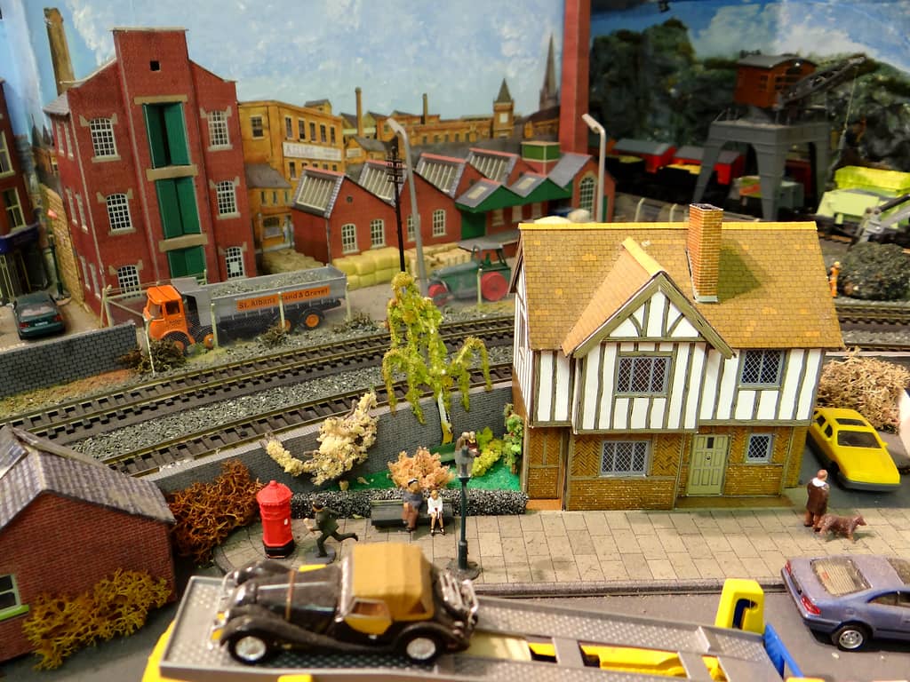 A model Tudor-style house with a small garden and pavement in front, a red pillar box and figures, a car transporter in the foreground, and a factory and builder’s yard beyond the railway.