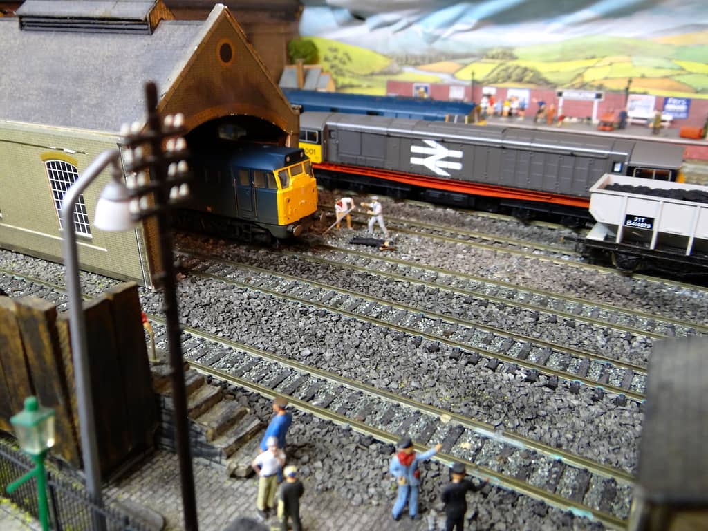 A blue diesel with a yellow front in the arched doorway of a brick engine shed, a grey diesel beside it, model workmen on the tracks and figures in the foreground.