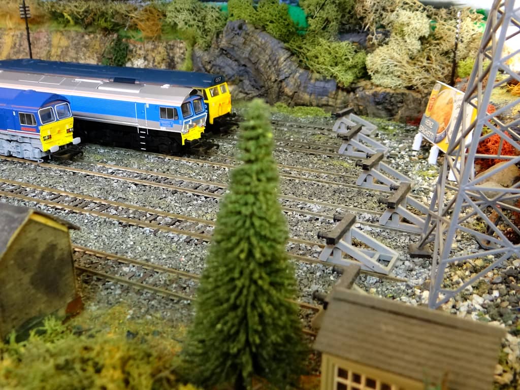 Three diesel locomotives with yellow fronts at the end of yard tracks below a rock face, beside buffer stops, a grey pylon, a billboard and a conifer.
